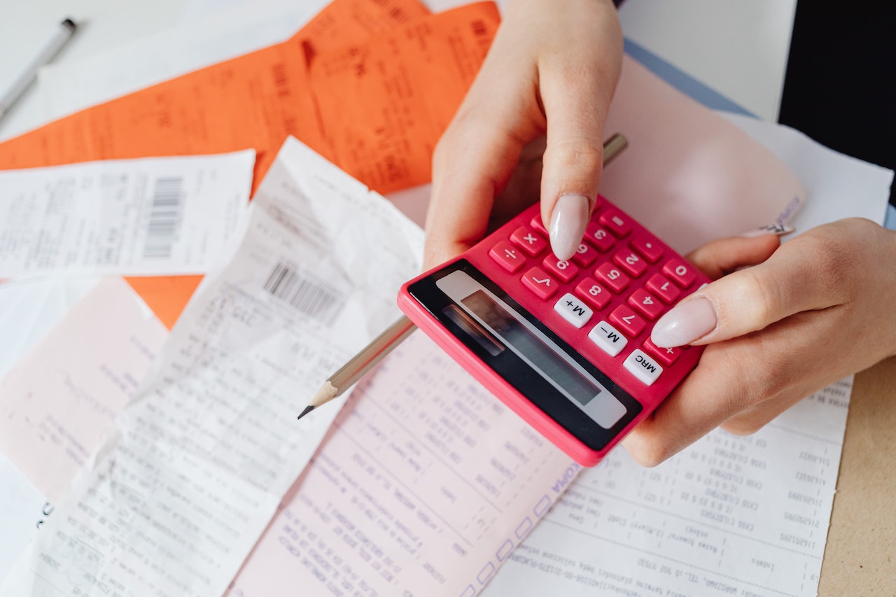 kobieta z różowym kalkulatorem w dłoniach stara się przygotować księgę przychodów i rozchodów na podstawie leżących na blacie dokumentów